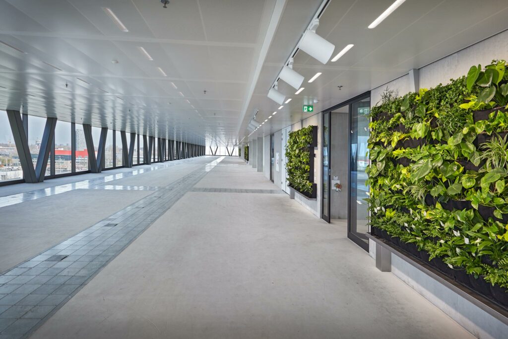 Lichte, moderne overdekte loopbrug aan Joan Muyskenweg met groene plantenwand en grote ramen met stadszicht.